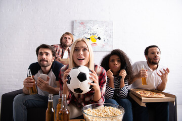 worried woman holding soccer ball while watching football championship with tense multiethnic...
