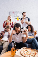 upset multicultural friends watching football championship near pizza and beer on blurred foreground