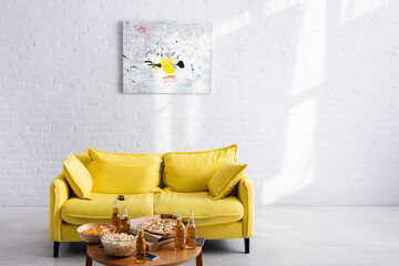 spacious living room with yellow sofa, picture on white wall, table with delicious pizza, beer, chips and popcorn in bowls