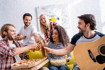 young man holding acoustic guitar while clinking bottles of beer with multiethnic friends
