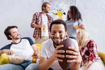 smiling african american man holding rugby ball near cheerful multicultural friends with painted faces on blurred background