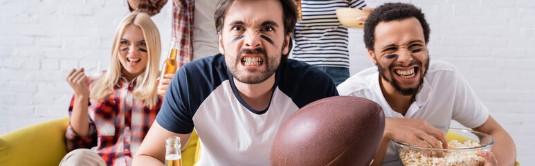 young man holding rugby ball and grimacing while watching championship near multiethnic friends,...