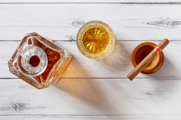 View of a crystal glass and decanter full of golden whisky, and cigar, shot from above on a distressed white wood background