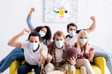excited multicultural friends in medical masks showing win gesture while watching rugby championship