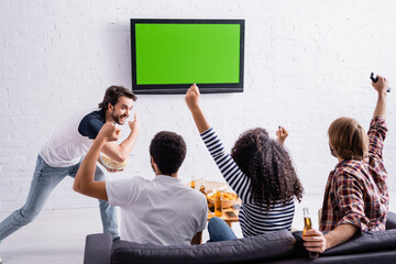 excited man pointing at lcd tv on wall near multicultural friends watching sport competition
