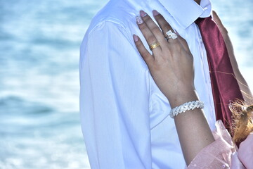 Newlyweds bride and groom near the sea. The bride's hand with a ring and bracelet lies on the shoulder of a man in a shirt. No faces.