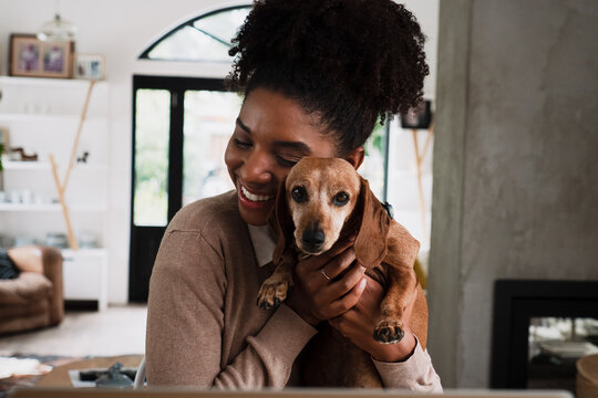 African American female smiling while snuggling cute puppy sitting at desk in modern kitchen.