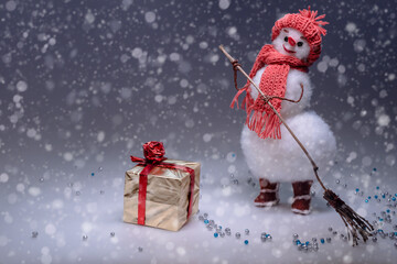 A snowman in a hat and scarf sweeps the snow and looks at the golden gifted box. Smooth Black...