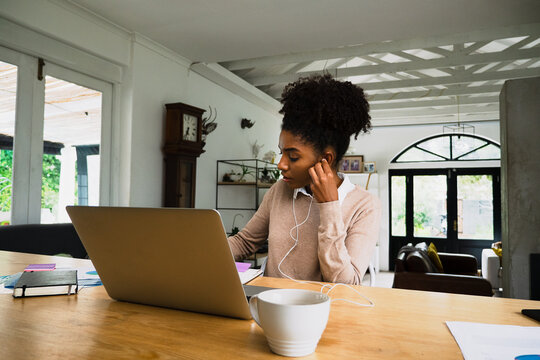 Busy Mixed Race Working Woman Writing Down Paperwork Wearing Earphones Listening To Video Call On Laptop Sitting At Desk In Modern Kitchen.