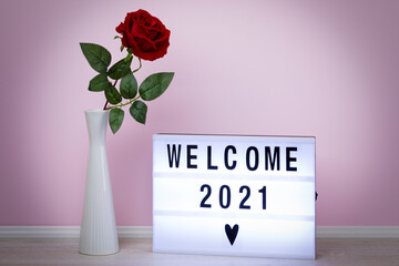 Welcome 2021, message written on lightbox, with rose in vase in pink room