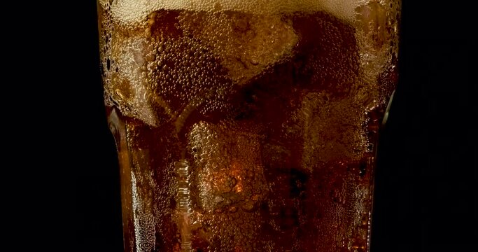 Truck shot of pouring carbonated drinks bubbles with ice cubes. Pouring soft drinks with ice and bubbles in glass on black background.