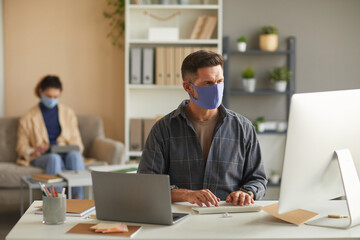 Fototapeta na wymiar Businessman in protective mask working on computer at his table with his colleague in the background at office