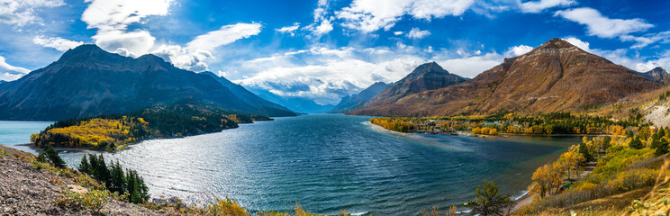 Upper Waterton Lake lakeshore in autumn foliage season sunny day morning. Blue sky, white clouds over mountains in the background. Landmarks in Waterton Lakes National Park, Alberta, Canada.