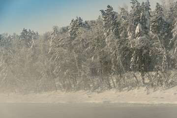 Frosty fog over winter river with snow and forest on bank. First ice on lake on cold day.