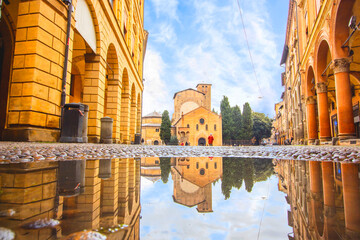Bologna in the morning, looking Saint Stefano church in square with sunlight and reflection in a puddle on the street. Elderly granmother walking - obrazy, fototapety, plakaty
