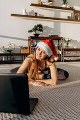 Girl trainer in a Santa Claus hat sits on a twine in front of a laptop screen in a yoga studio.New year's eve workout concept.Online stretching and yoga training in the coronavirus pandemic.