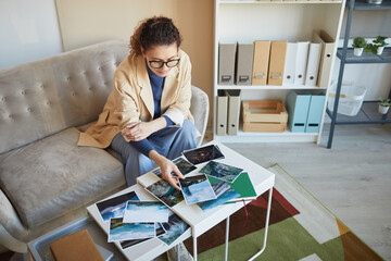 Young woman in eyeglasses sitting on sofa at the table and choosing the best photos for design project