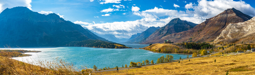 Naklejka na ściany i meble Middle Waterton Lake lakeshore in autumn foliage season sunny day morning. Blue sky, white clouds over mountains in the background. Landmarks in Waterton Lakes National Park, Alberta, Canada.