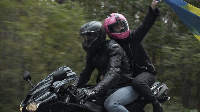Young interracial couple in helmets riding motorbike with woman holding Ukrainian flag. Middle Eastern man and Caucasian woman racing with national symbol. Pride and lifestyle concept.