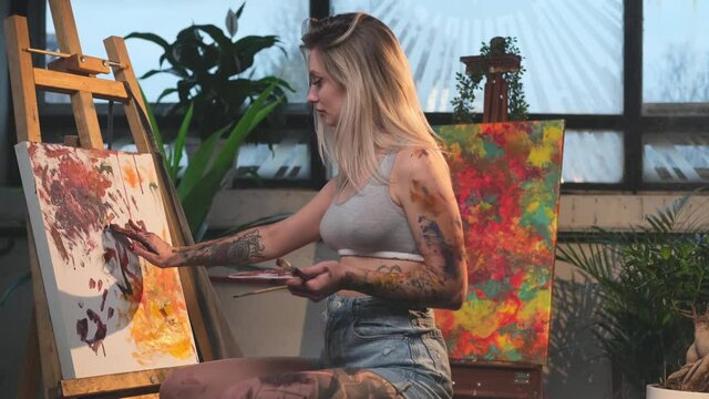 Seductive blond haired female painter with tattoos in short clothing drawing on canvas with her arm with paints on palette in warm and tranquil workplace with bamboo furniture and another abstract