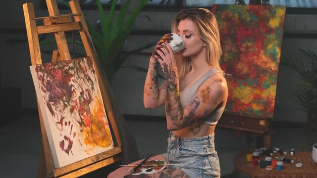 Grimy and glamour female artist with blond hairs poses in spacious and warm creative studio sitting on chair and drinking coffee around canvas and artwork with table there are many different paints.