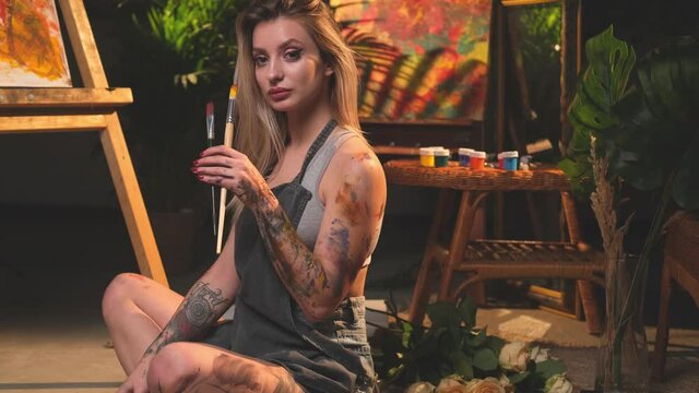 Grimy charming female painter with apron and blond hairs poses sitting on floor and holding two paintbrushes in dark and atmospheric apartment there are canvas and artwork with bamboo furniture.