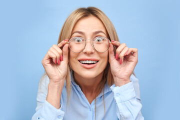 OMG! Wow! Studio shot blonde girl with happy smiling face holds hands on the rim of glasses, looking at the camera with curiosity, emotional reaction to amazing news isolated on blue background