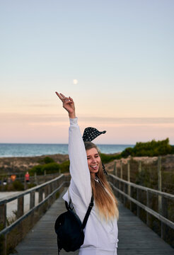 Side view of positive female tourist showing tongue and v sign while standing with closed eyes on wooden embankment at seaside and enjoying freedom during summer vacation