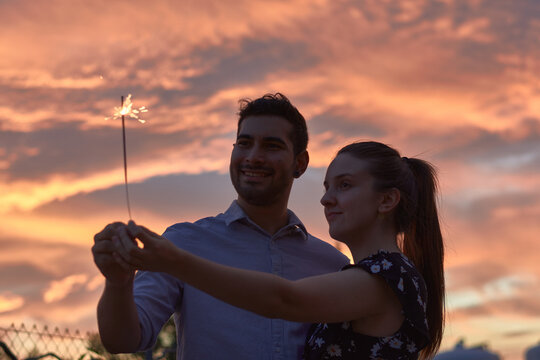 Low angle of tranquil couple embracing and standing with sparkler on background of sunset in summer