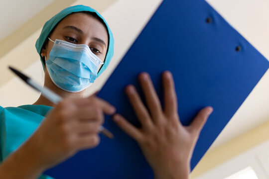 Concentrated young nurse in blue uniform and mask making medical notes on clipboard while working in hospital