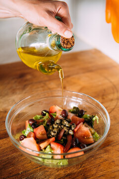 Crop anonymous housewife adding olive oil into bowl with fresh healthy vegetable salad while preparing lunch in kitchen
