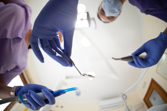 Blurred female dentist with mirror and drill and assistant with suction device standing against clinic ceiling during curing procedure