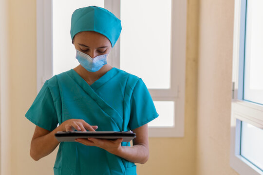 Young Nurse In Blue Scrubs And Medical Mask Reading Information On Tablet While Standing In Corridor Of Modern Hospital