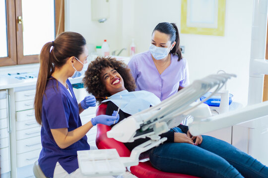 Female assistant looking to woman while dentist holding instrument to curing teeth of African American lady in clinic