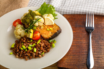 Baked fish with broccoli and lentils in a white plate with rosemary and lemon on a napkin and a round stand next to a fork.