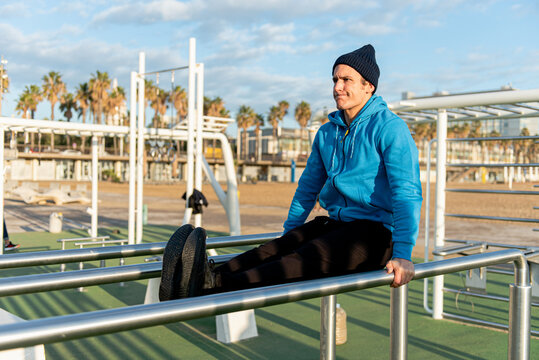 Full body of determined adult ethnic male in sportswear doing exercise for abdominal muscles on parallel bars during outdoor fitness workout on sports ground