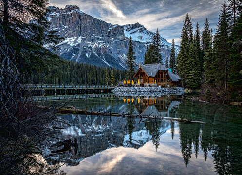 Magnificent scenery of Emerald Lake with bridge over water and wooden house on shore surrounded by green pine forest against rocky mountains covered with snow in Yoho National Park in Canada