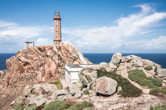 Picturesque scenery with old lighthouse tower and houses located on rough rocky cliff near sea on Cape Vilan peninsula on Spanish coast against cloudy blue sky in summer day
