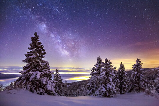 Glowing Northern lights and Milky Way in night sky over mountainous terrain with coniferous trees covered with snow in wintertime