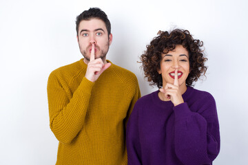 Smiling Young couple wearing knitted sweater standing against white wall makes shush gesture, holds...