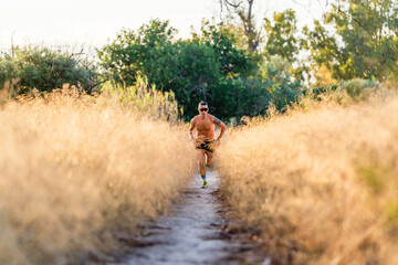 Full body shirtless male athlete with sunglasses running on narrow footpath among tall grass during outdoor workout in nature