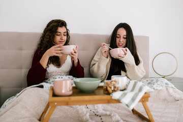 two white women in a bed having breakfast. They wear lingerie and a cardigan
