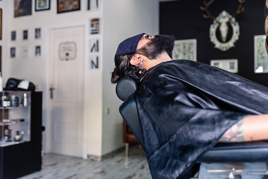 Side view of bearded hipster male client with towel on face sitting in comfortable chair and relaxing during procedure in modern barbershop