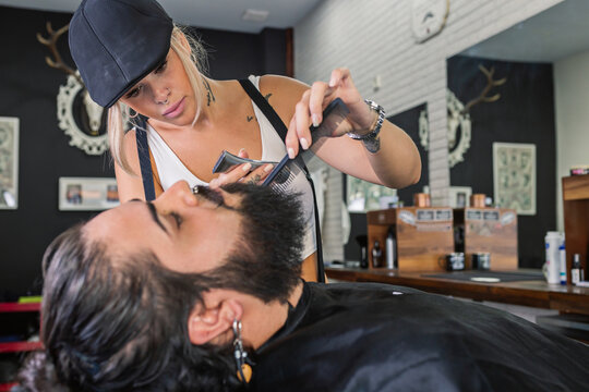 Side view of stylish tattooed female master shaving bearded customer with electric machine while working in professional barbershop