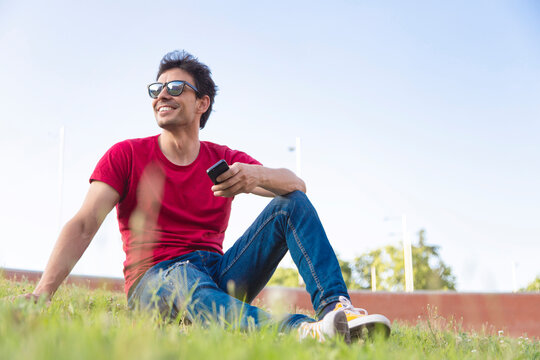 Attractive young man dressed with red t-shirt smiling in the park with sunglasses and smartphone