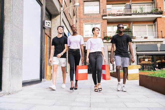 Low angle full length of group of cheerful multiracial millennial friends in trendy clothes carrying shopping bags and walking on urban street while enjoying shopping time together