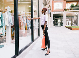 Full body side view of young slim African American female shopper in stylish outfit with shopping bag in hand standing near window of modern clothing store and looking at new collection