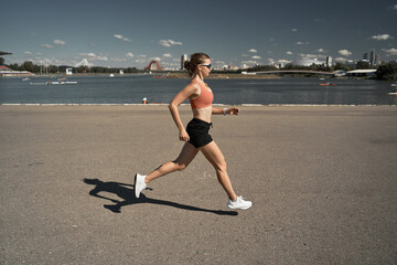 Side view full body of energetic motivated fit female in activewear with fitness tracker jogging on paved embankment near river in city