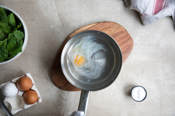Top view of metal saucepan with boiling water and egg during poached eggs preparation on kitchen table with ingredients