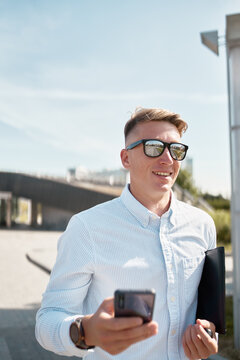 Confident young male in casual shirt and trendy sunglasses carrying paper folder walking on urban street and using mobile phone